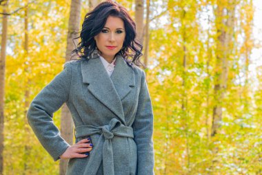 Şık ceketli genç bir esmer kız, sonbahar sarı yapraklarının arka planında parkta yürüyor.