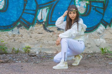 Beyaz eşofmanlı genç bir kız onun kalçalarına oturur, bir beyzbol şapkasını düzeltir ve bir çizimle bir duvarın arka planındaki kameraya bakar.