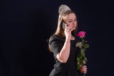 Kafasında çapı olan ve elinde bir gül çiçeği olan bir prenses suretinde genç bir kız koyu bir arka planda duruyor.