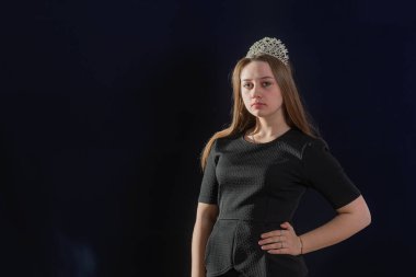 Kafasında çapı olan bir prenses suretinde genç bir kız karanlık bir arkaplanda duruyor ve kameraya bakıyor, eli belinde.