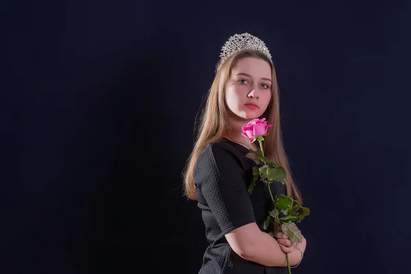 A young girl in the image of a princess with a diadem on her head and a rose flower in her hands stands on a dark background and looks into the camera