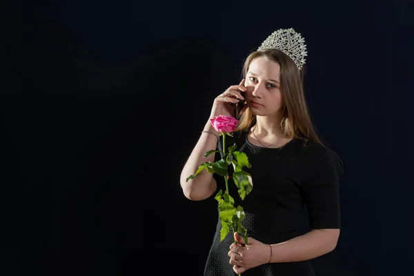 Young girl in the image of a princess with a diadem on her head is standing against a dark background and talking on the phone. Looks into the camera