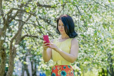 Sarı elbiseli genç bir kadın çiçek açan bir elma bahçesinin ortasında duruyor ve cep telefonuyla kendi fotoğrafını çekiyor.