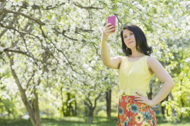 Parlak sarı-kırmızı bir yaz elbisesi içindeki genç bir kız, arka planda çiçek açan bir elma bahçesinde akıllı bir telefonla kendi fotoğraflarını çekiyor.