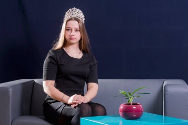 Genç bir kız, esmer, kanepede oturuyor. Kafasına diyadem takmış. Elleri dizlerinin üstüne çökmüş. Kameraya bak..