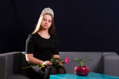 Genç bir kız, esmer, kanepede oturuyor. Kafasına diyadem takmış. Elinde bir gül çiçeği tutuyor. Kameraya bak..