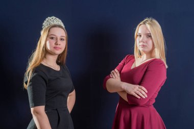 İki genç kız karanlık bir arkaplanda yan yana duruyor. Kameraya bakıyorlar. Birinin kafasına diyadem takılmış..