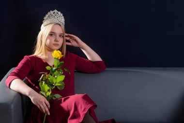 Genç bir kız, esmer, kanepede oturuyor. Kafasına diyadem takmış. Elinde sarı bir gül tutuyor. Kameraya bak..