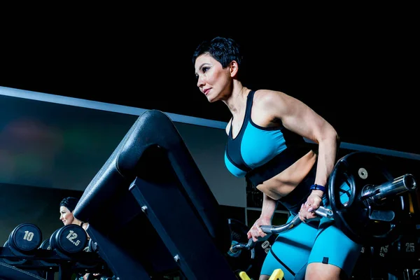 A slender middle-aged woman in a sports suit trains with a barbell in a gym. A charge of vivacity and spirit for the whole working day.