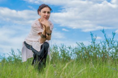 Young woman performs yoga exercises. Stands on a green field against the blue summer sky. Healthy lifestyle.