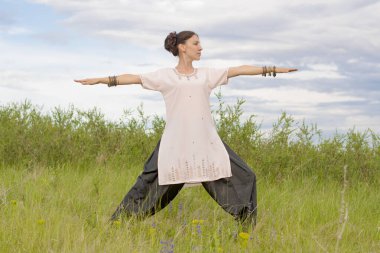 Young woman performs yoga exercises. Stands on a green field against the blue summer sky. Healthy lifestyle.