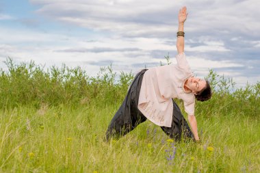 Young woman performs yoga exercises. Stands on a green field against the blue summer sky. Healthy lifestyle.