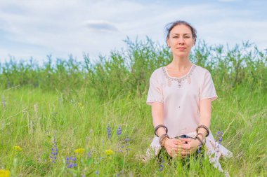 Genç bir kadın, lotus pozisyonunda yeşil bir çimenlikte otururken meditasyon yapıyor. Yoga dersi açık havada..