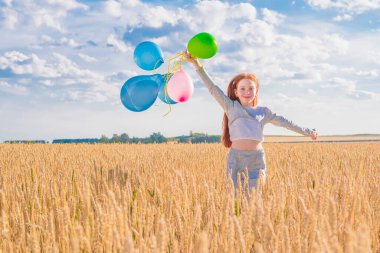 Elinde renkli balonlarla genç, mutlu, kızıl saçlı kız mavi bulutlu bir gökyüzüne doğru sarı buğday tarlasında koşuyor. Kameraya bakıyor. Yakın plan..