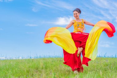 Parlak ulusal bir kostüm giymiş genç bir kadın yeşil bir çayırda hayranlarıyla mavi bir yaz gökyüzüne karşı oryantal bir dans ediyor. Keyifli zaman geçirme.