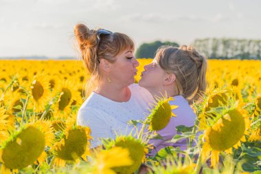 Anne ve kızı sarı bir ayçiçeği tarlasının ortasında durur ve bir öpücükle birbirlerine uzanırlar. Ebeveyn ve çocuk arasındaki ilişki. Pozitif duygular.