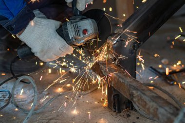 Kesilmiş bir işçinin elleri metal bir yapının kaynak dikişlerini temizlemek için öğütücü kullanır. Sıcak metal kıvılcımları uçuşuyor.