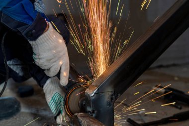 Kesilmiş bir işçinin elleri metal bir yapının kaynak dikişlerini temizlemek için öğütücü kullanır. Sıcak metal kıvılcımları uçuşuyor.