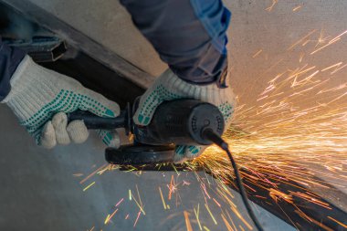 Kesilmiş bir işçinin elleri metal bir yapının kaynak dikişlerini temizlemek için öğütücü kullanır. Sıcak metal kıvılcımları uçuşuyor.