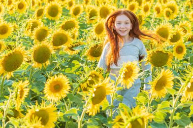 Genç kızıl saçlı bir kız sarı bir ayçiçeği tarlasında koşar, gülümser ve kameraya bakar.