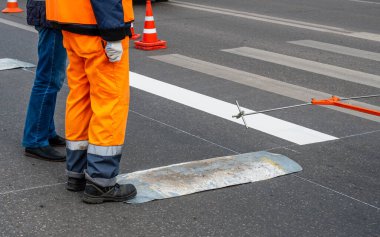 İki özel giysili işçi bir yaya geçidinin işaretlendiği şehir yolunun kenarında duruyor..