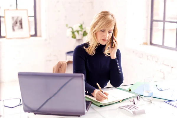 一位自信的女商人坐在手提电脑后面工作时 一边打电话谈生意 总部办公室 — 图库照片