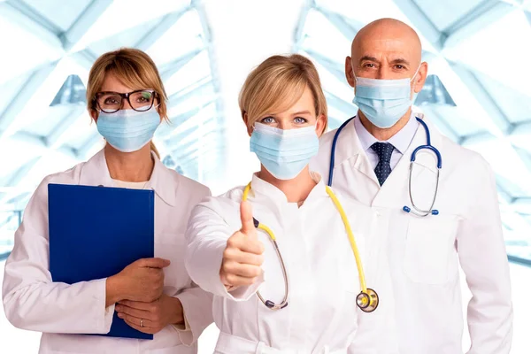 顔のマスクを着用した医療チームのショットと一緒に病院のホワイエに立っている間 女性医師は親指を放棄 — ストック写真