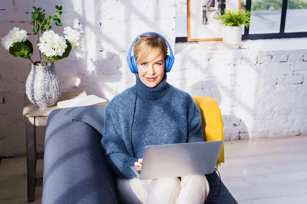 소파에 앉아서 노트북을 사용하면서 헤드폰을 아름다운 미소짓는 여성의 초상화 — 스톡 사진