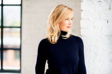 Boğazlı kazak giyen ve pencerenin yanında kameraya bakan gülümseyen bir kadının portresi.. 