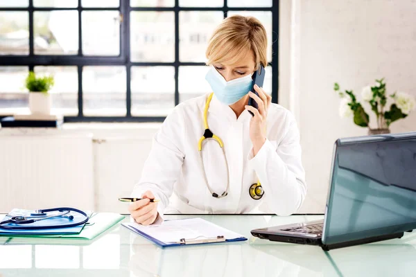 사무실에 앉아서 환자와 휴대폰으로 통화하는 의사의 마스크를 — 스톡 사진