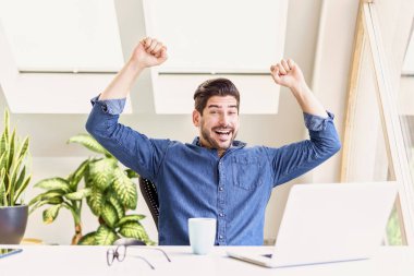 Neşeli iş adamı, evinde masa başında oturup dizüstü bilgisayarla çalışırken kollarını havaya kaldırarak kutlama yapıyor. Merkez Ofis.