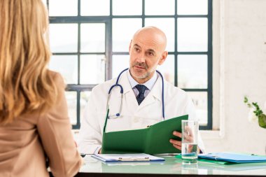 Orta yaşlı erkek doktorun, doktorların odasında masasında otururken hastasıyla konuşması.. 