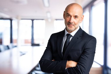 Toplantı odasında kolları çapraz dururken takım elbiseli ve kravatlı bir yöneticinin portresi..