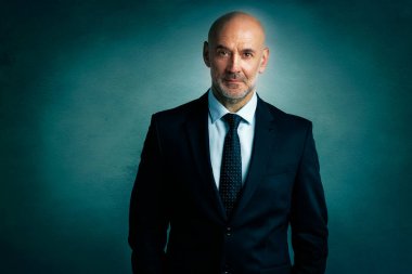 Tecrit edilmiş karanlık arka planda dururken takım elbiseli ve kravatlı kıdemli işadamının stüdyo portresi.