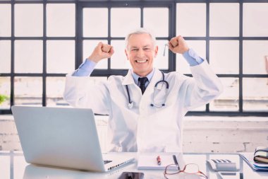 Doktorun muayenehanesinde laptopun önünde otururken başarısını kutlayan başarılı erkek doktor..