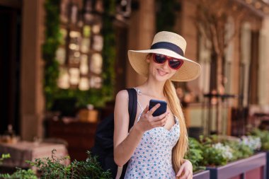 Saman şapka takan ve sokakta yürürken cep telefonuyla mesajlaşan gülümseyen genç bir kadının portresi..