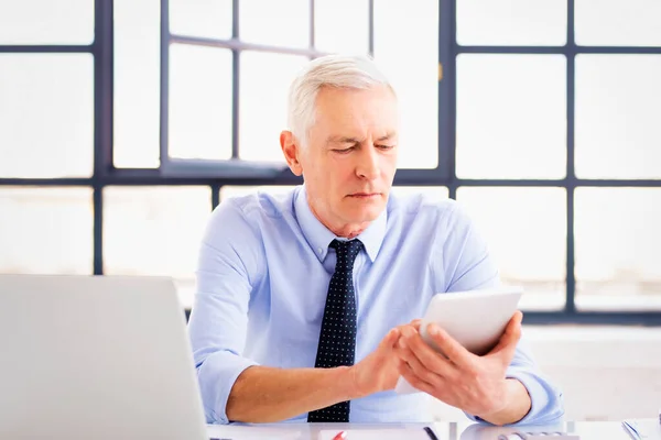 デスクに座ってオフィスで働いている間 彼の手にデジタルタブレットを保持している上級財務コンサルタントのビジネスマンのショット — ストック写真