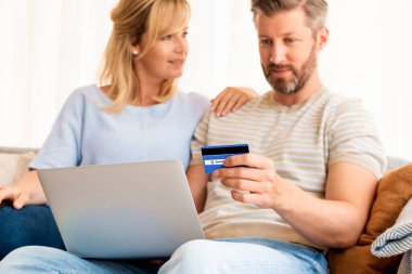 Evden online alışveriş. Karısı yanında oturmuş internetten alışveriş yaparken elinde banka kartı tutan yakışıklı bir adam.. 