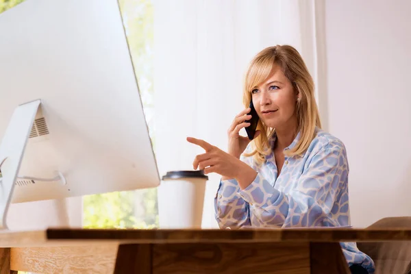 特写镜头拍摄的是一位快乐的金发女人 一边坐在办公桌前 一边在家工作 一边打电话和使用电脑 总部办公室 — 图库照片
