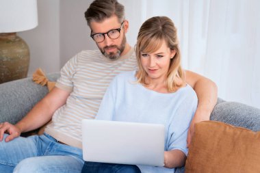 Evdeki kanepede dinlenen ve internette gezinirken dizüstü bilgisayar kullanan sevimli bir çiftin fotoğrafı..