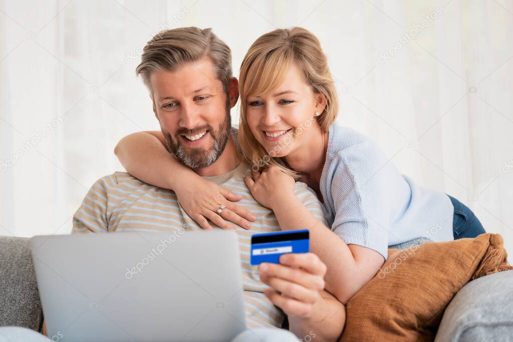 Online shopping from home. Handsome man holding bank card in his hand while her wife sitting next to him and using laptop while shopping online. 