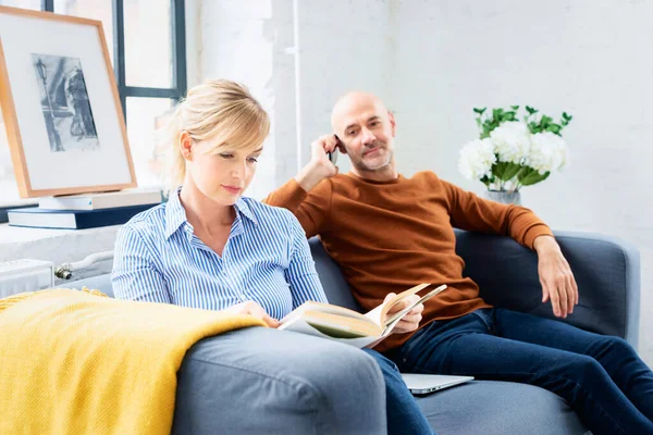 Mujer Encantadora Leyendo Libro Mientras Marido Sentado Lado Sofá Hablando —  Fotos de Stock