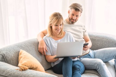 Mutlu çift evdeki kanepede oturuyor ve internette gezinirken dizüstü bilgisayar kullanıyorlar. Elinde cep telefonu tutan yakışıklı bir adam..