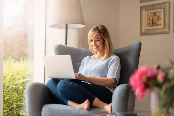 Blond Haired Smiling Woman Using Her Laptop While Sitting Armchair — Stock Photo, Image