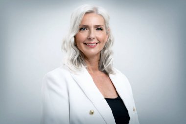 Gri saçlı iş kadınının stüdyo portresi. Kameraya bakıyor ve gri arka planda dikilirken gülümsüyor. Boşluğu kopyala. 