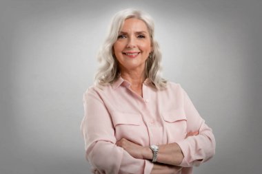 Gri arka planda kolları çapraz duran gri saçlı yaşlı kadının stüdyo portresi. Boşluğu kopyala. 