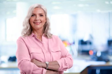 Gri saçlı kıdemli iş kadını kameraya bakıyor ve ofiste dururken gülümsüyor..