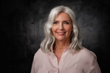 Stüdyo portresi, gri saçlı yaşlı kadının kameraya bakıp izole edilmiş karanlık arka planda dururken gülümsemesi. Boşluğu kopyala.