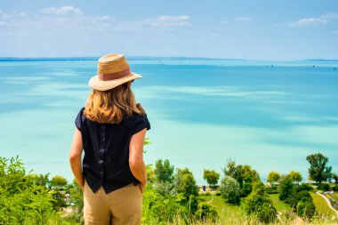 Dışarıda dikilip deniz manzarasına bakarken hasır şapka takan bir kadın resmi. Hayal kurmak.