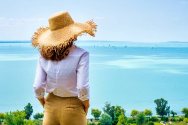 Dışarıda durup deniz manzarasına bakarken hasır şapka ve beyaz gömlek giyen bir kadın. Hayal kurmak.